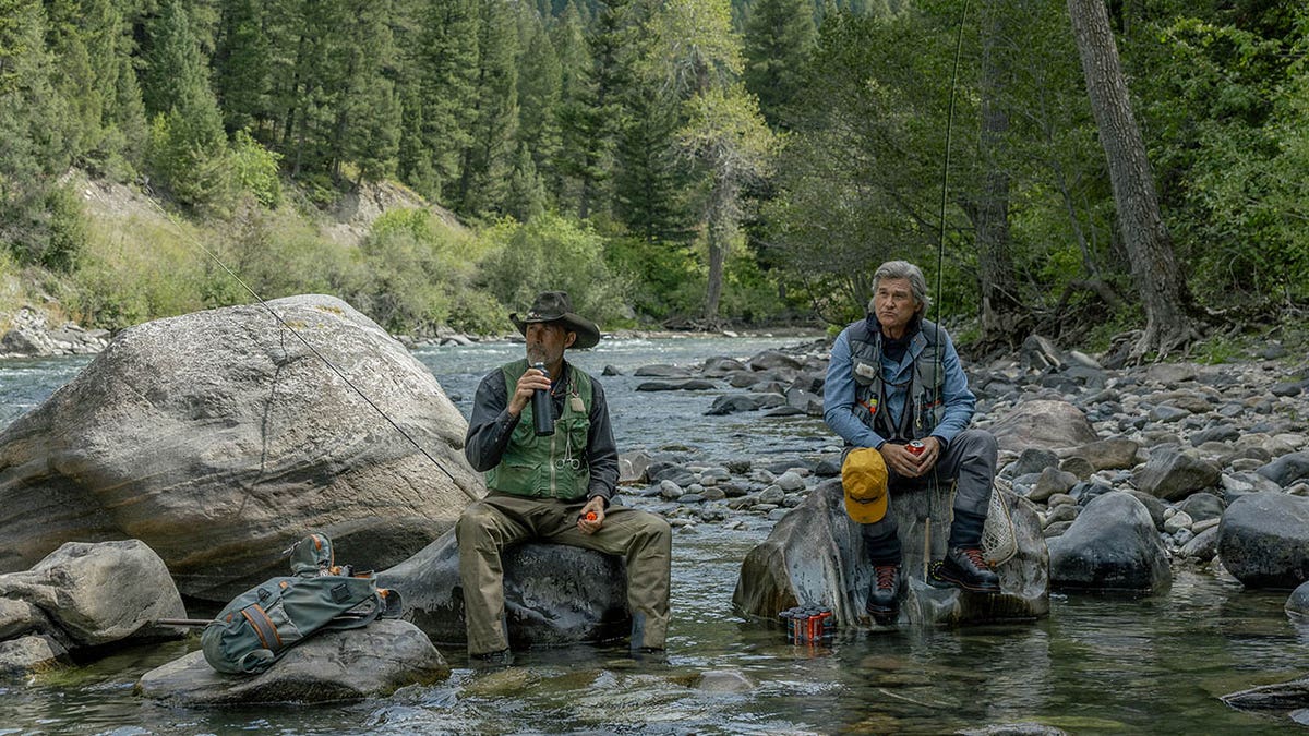 Matthew Fox and Kurt Russell in fishing gear sitting on large rocks in a shallow river surrounded by forest.