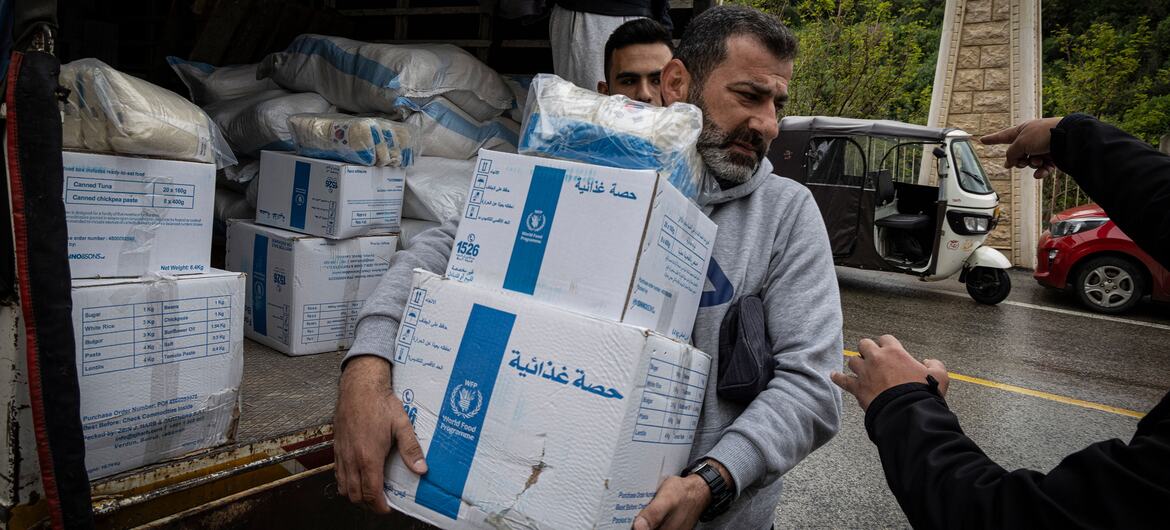 A man carries boxes of food aid from a truck. The boxes are labeled with the World Food Programme (WFP) logo and Arabic text. The scene takes place outdoors in a rural area of Lebanon, showing a humanitarian aid distribution to displaced families.