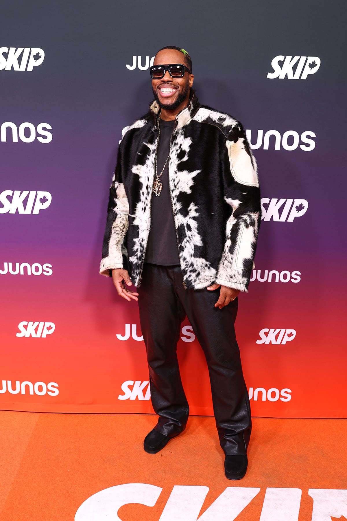 A man poses wearing a cow print jacket on the orange carpet with leather pants, oversized sunglasses and a large pendant necklace.