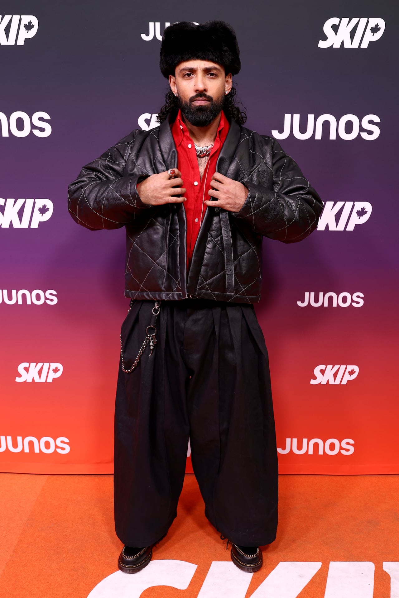 A man poses for photos in baggy black pants, a leather jacket and a fur hat.