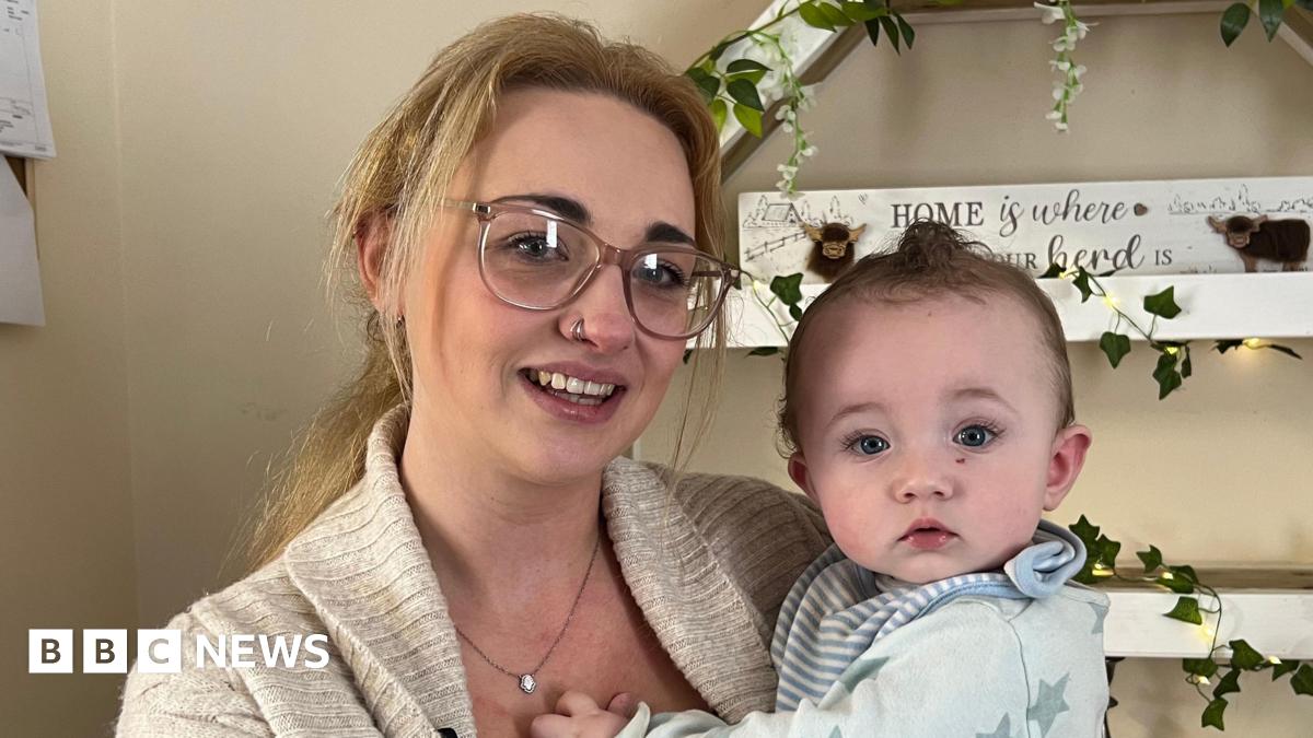 Kiely Luscombe pictured with blonde hair tied back in a pony tail. She is wearing glasses and a beige cardigan with a black top underneath. She is holding Arnold who is wearing a blue baby grow with stars on.