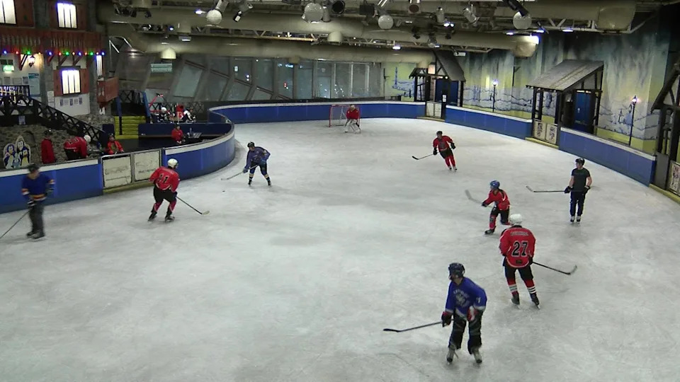 A number of ice hockey players playing on an undercover ice rink. They are all wearing helmets and carrying hockey sticks. They are split into a red team and a blue team.