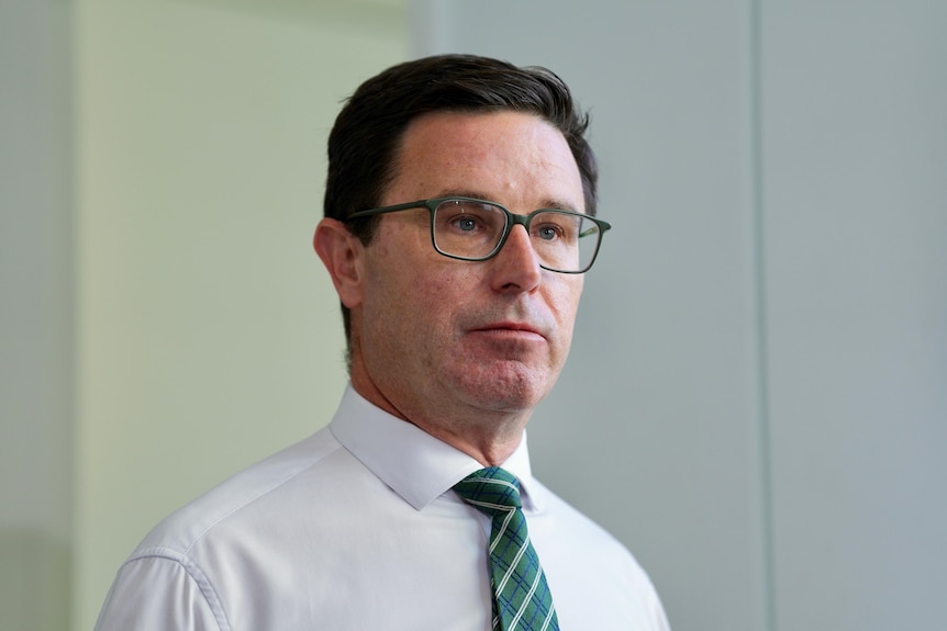 A white man with brown hair and glasses wearing a white shirt and green tie stares ahead 