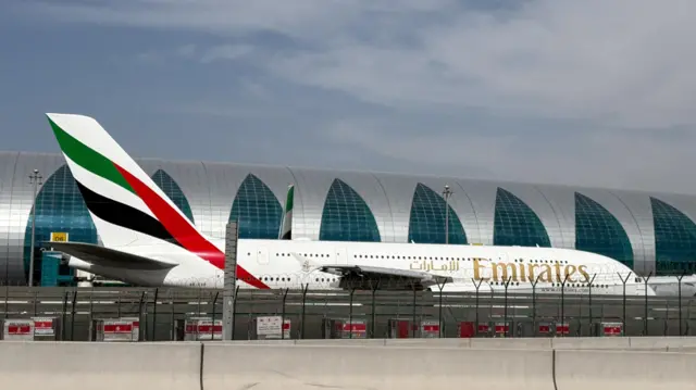 An Emirates airplane sits at the Dubai International Airport with a curved building in the background.