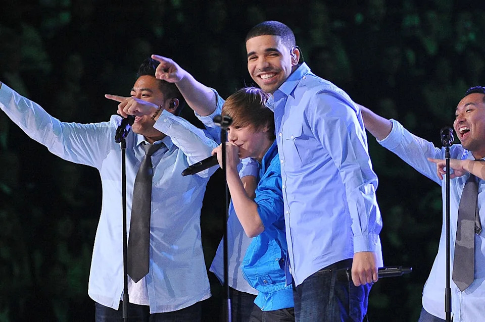 Group of musicians on stage, performing with energy. Central figure in a vibrant jacket, surrounded by others pointing excitedly. Audience visible