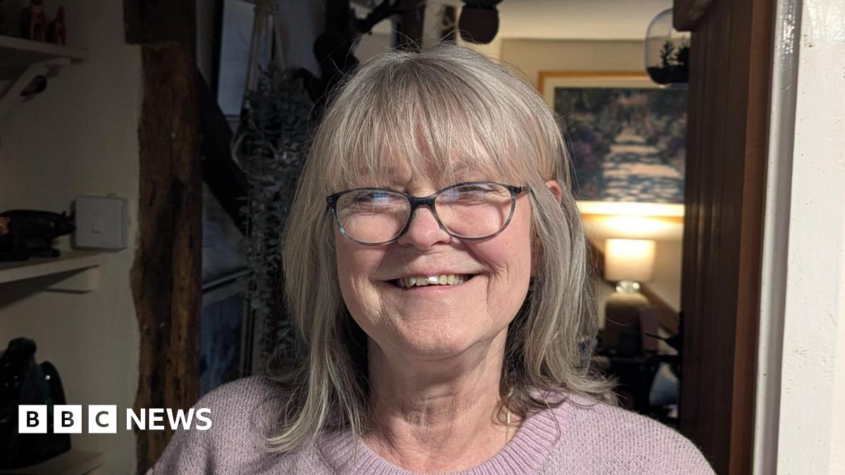 Jacky Smith smiles at the camera inside her home. She has short grey hair and wears glasses along with a light pink knitted jumper.