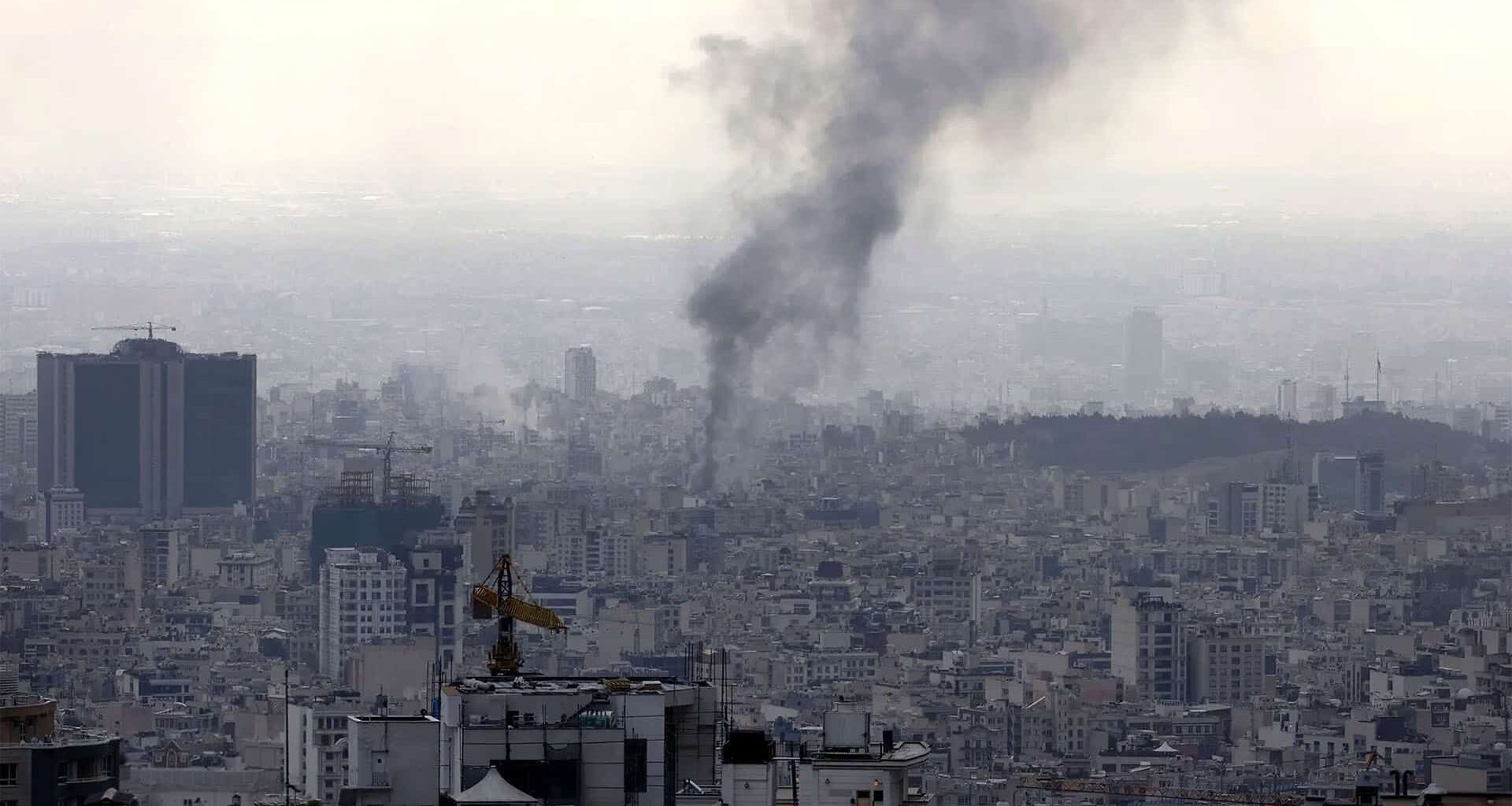 Panoramic view of Tehran as a thick column of black smoke rises from the city after an explosion, among dense buildings and construction cranes under a greyish sky.