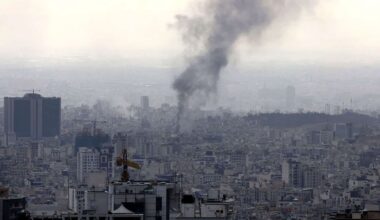 Panoramic view of Tehran as a thick column of black smoke rises from the city after an explosion, among dense buildings and construction cranes under a greyish sky.