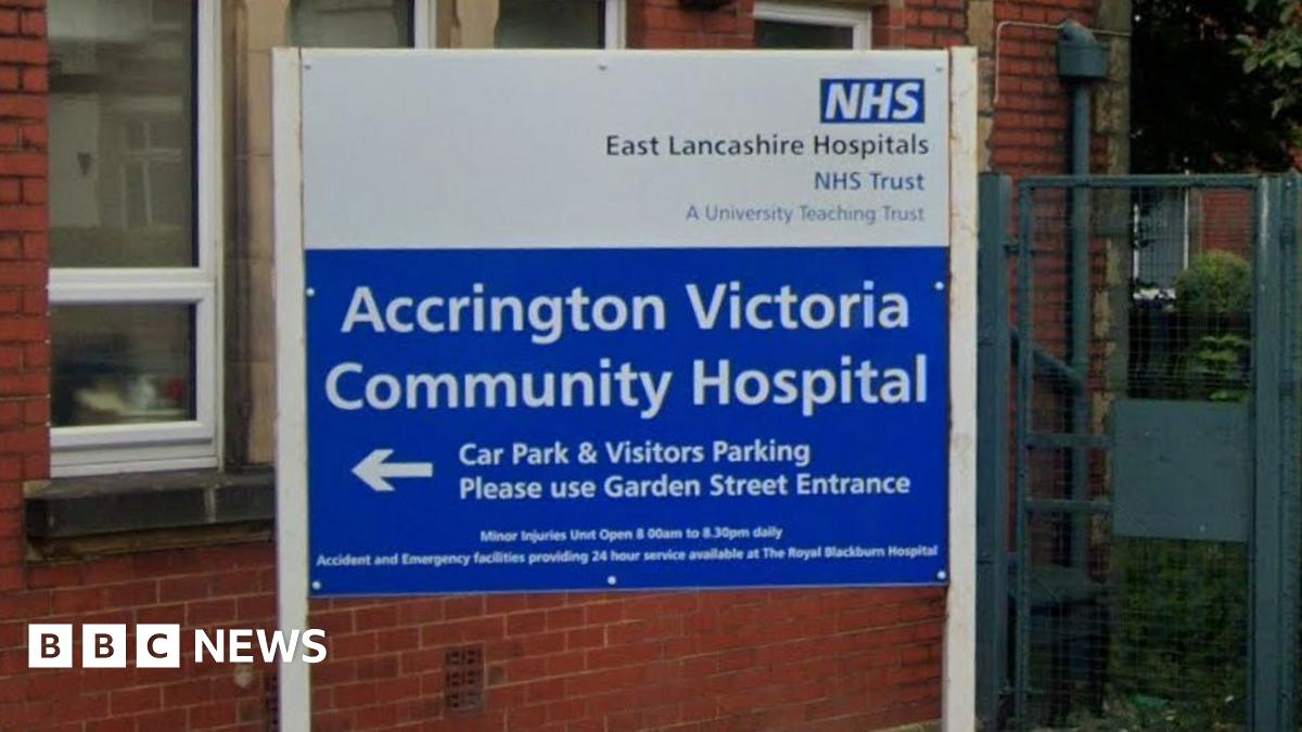 A street view image of Accrington Victoria Hospital, a red brick building with blue and white signs either side of a white awning with blue legs over the entrance and a man walking under it to the front door
