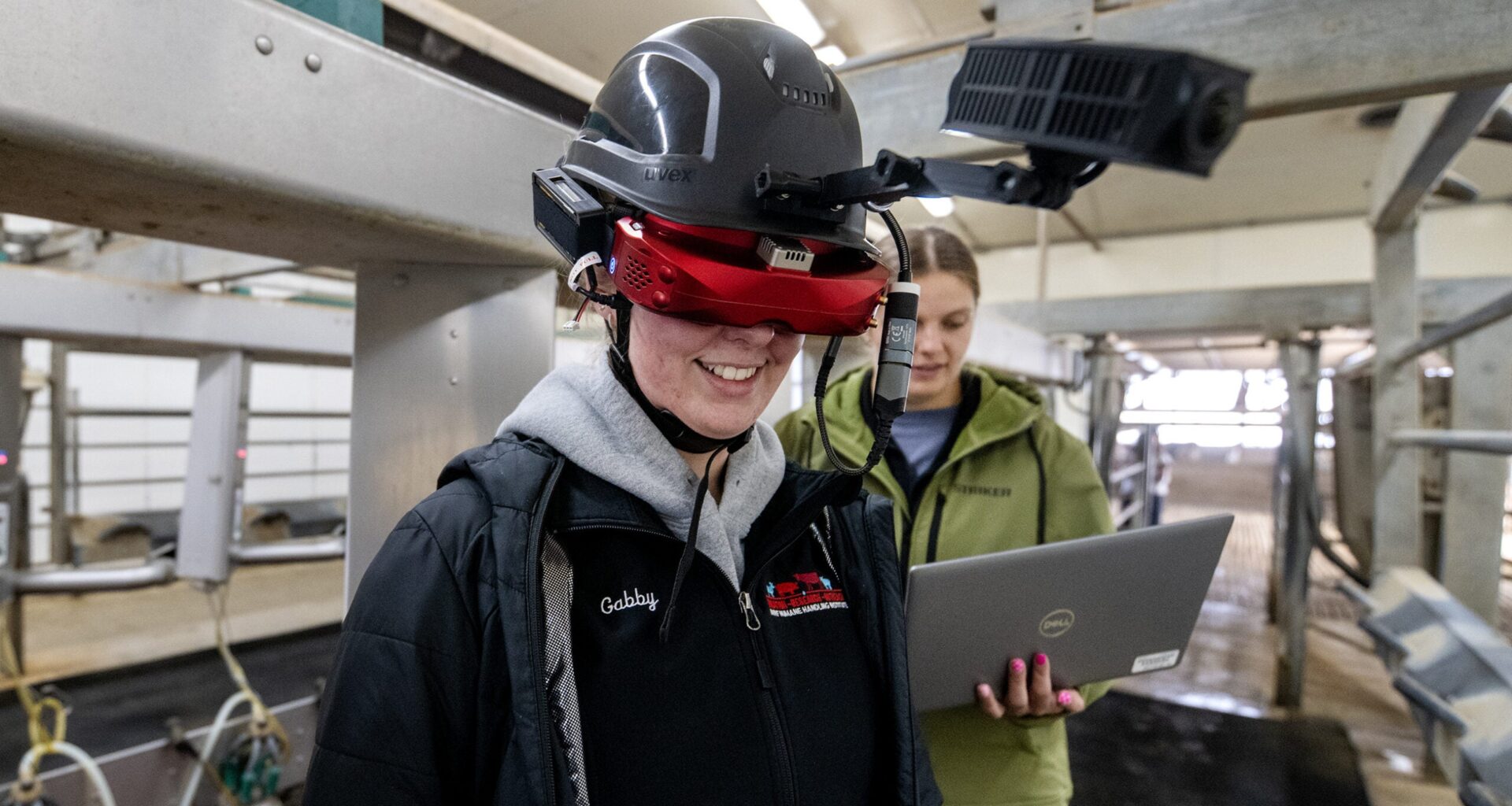 A person wearing advanced augmented reality equipment stands in a facility, while another person in the background works on a laptop.