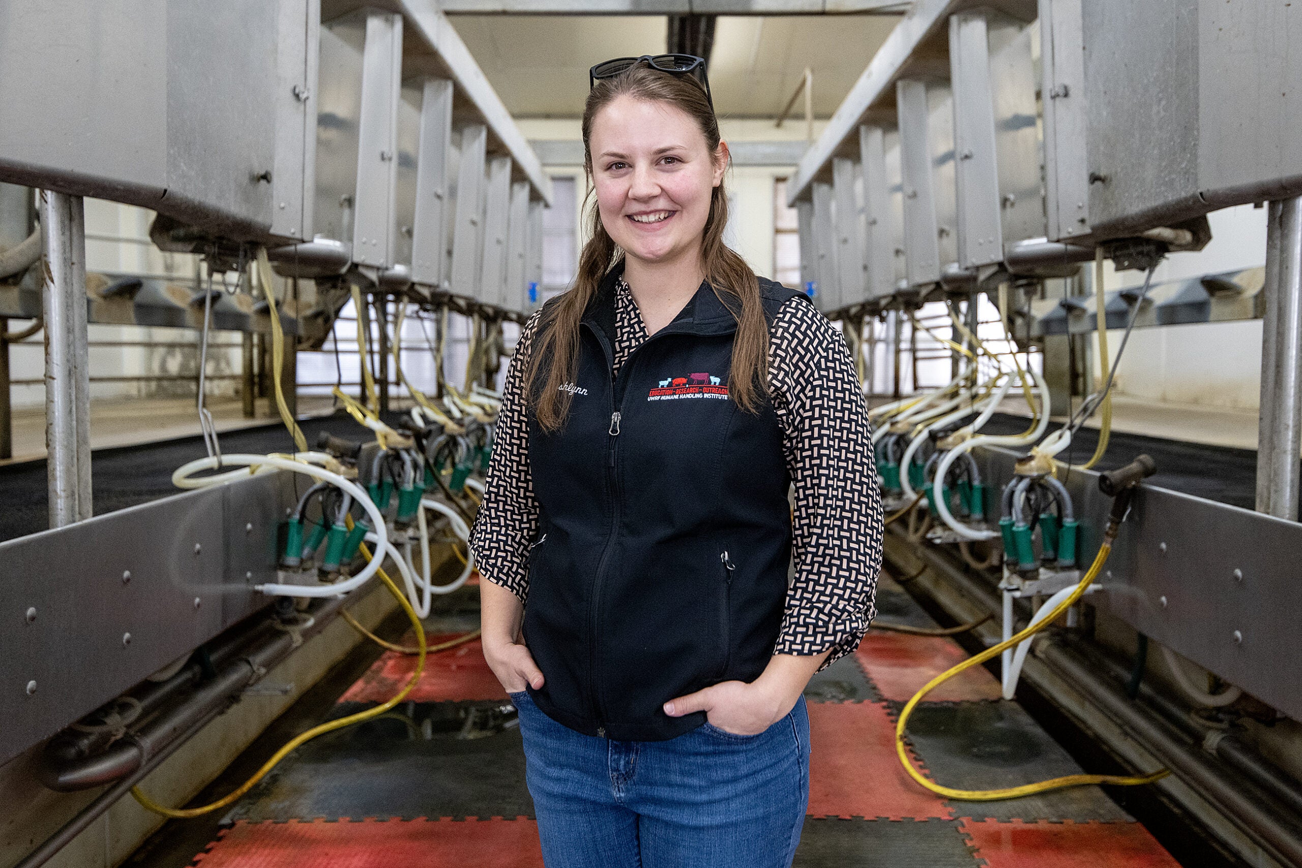 A woman stands smiling in a modern dairy farm milking parlor, surrounded by milking equipment and machinery.