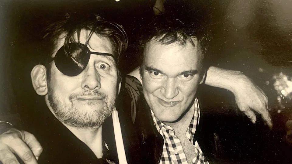 A black and white photograph of Shane MacGowan and film director Quentin Tarantino. Shane is wearing an eye patch and has his arm around the shoulder of Quentin - who in turn has his arm around Shane's shoulder. They are both looking into the camera. 