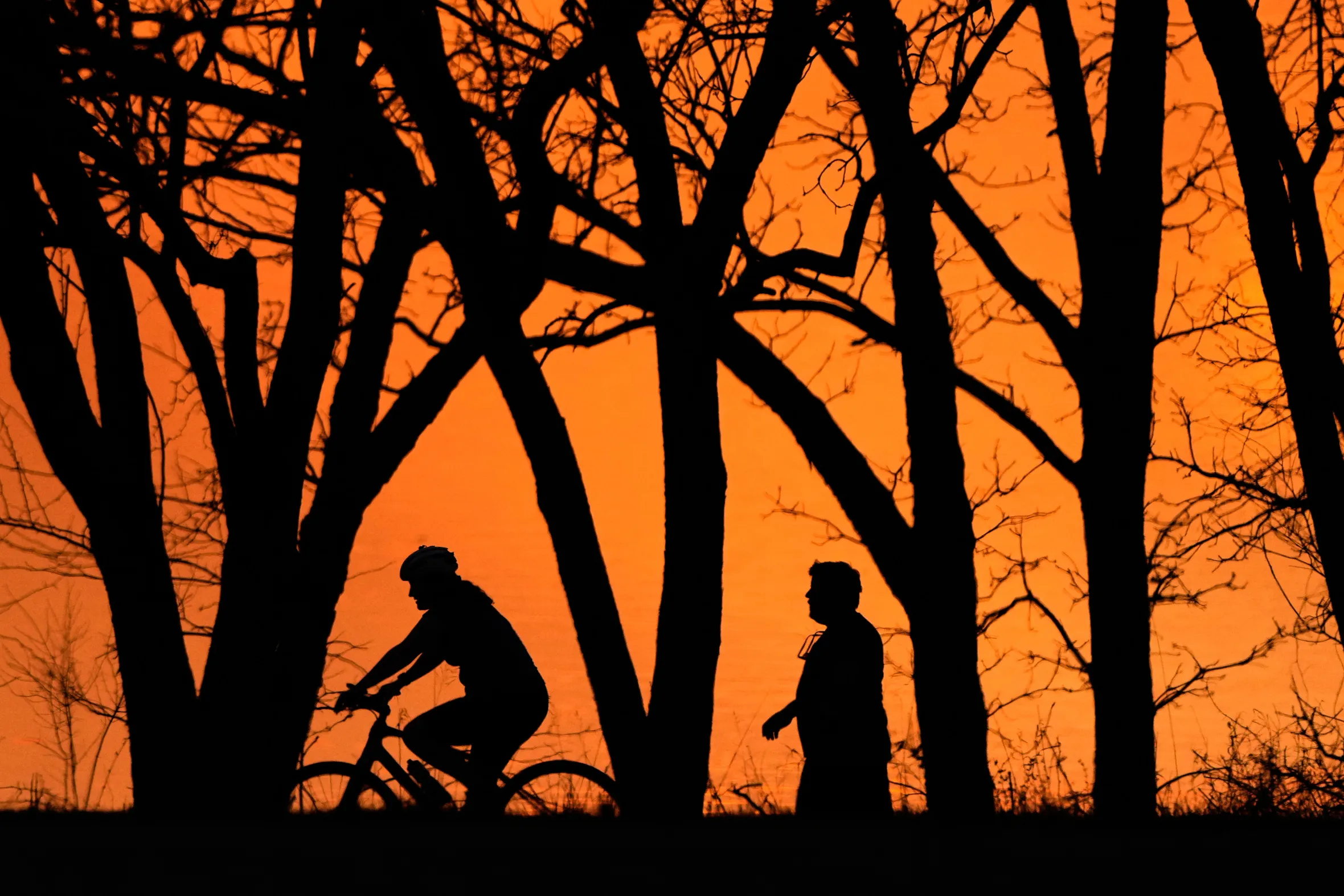 Silhouetted against a bright orange sky are trees, a person on a bicycle, and a person walking.