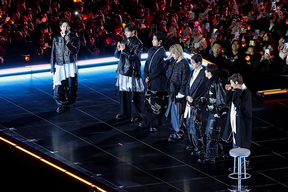BTS on stage, dressed in coordinated black and white outfits, with a large crowd of fans in the background.