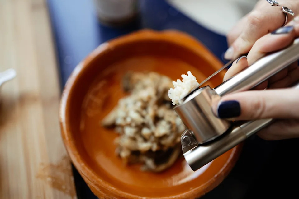 Close-up of hands pressing garlic into a ceramic bowl containing mushrooms on a wooden table