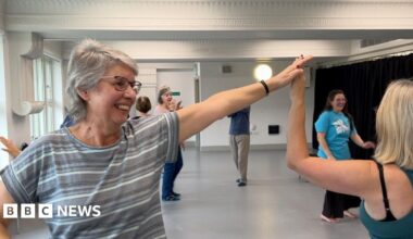 A woman with grey shoulder length hair and a striped top has her left arm outstretched and raised dancing with her partner, whose face isn't visible. A handful of other dancers and an instructor is visible in the background.