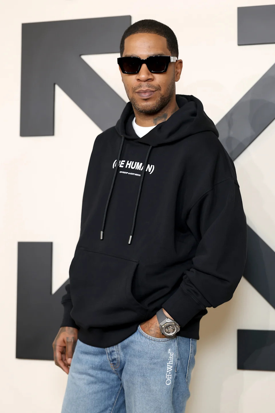 Person in casual style with sunglasses and a black hoodie featuring text, posing in front of a large logo backdrop