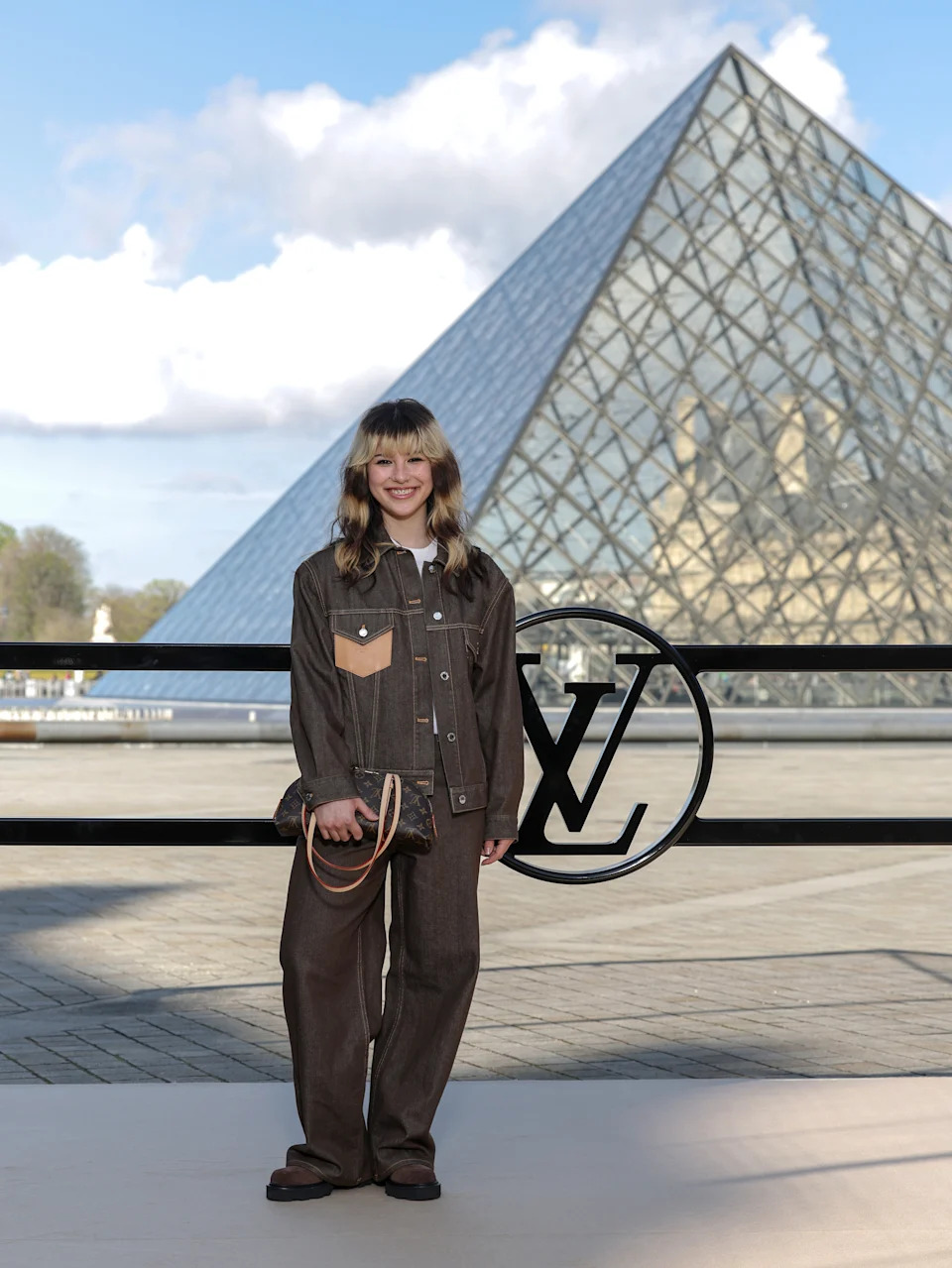 Person in a chic, oversized brown pantsuit poses in front of a glass pyramid, likely at a fashion event