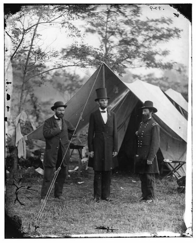 Three men stand in front of a canvas tent outdoors. The man in the center, tall and wearing a suit and top hat, is flanked by two men in hats, one in a military uniform. Trees and camp equipment are visible around the tent.