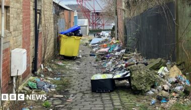 Rubbish filling an alleyway. Full bins, a suitcase, pillows, and litter are strewn across the length of the path.