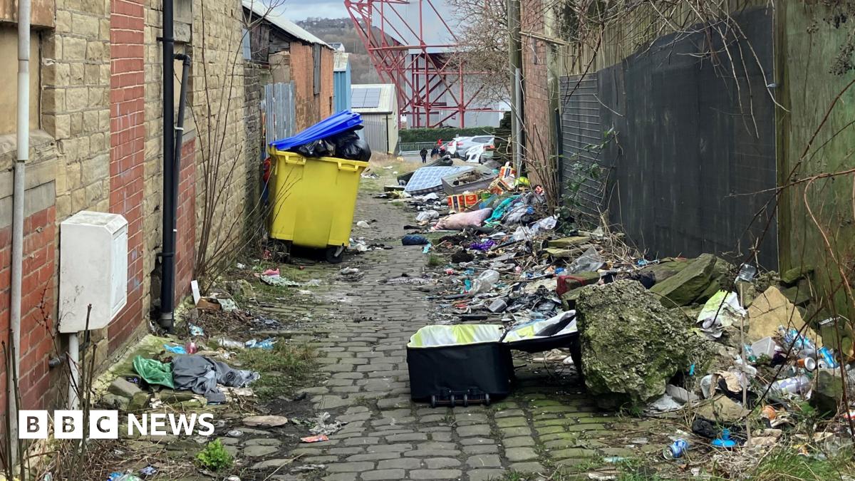 Rubbish filling an alleyway. Full bins, a suitcase, pillows, and litter are strewn across the length of the path.