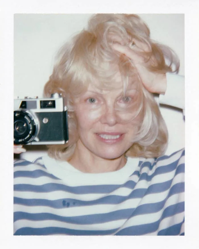 Person smiling, holding a vintage camera near their face, wearing a striped shirt