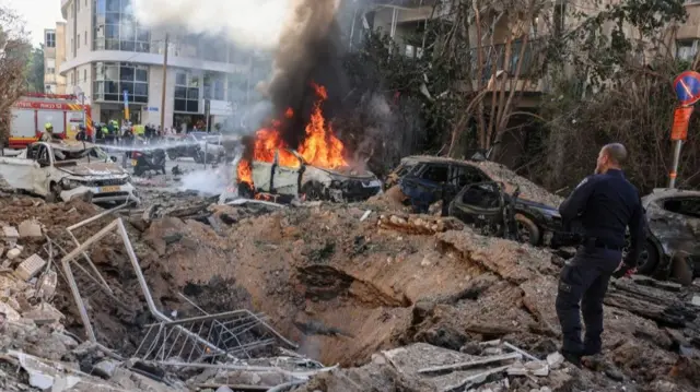 A man in uniform walks around the edge of a large crater in the middle of a street. Several burned-out cars are beside the crater, one of which is still on fire.