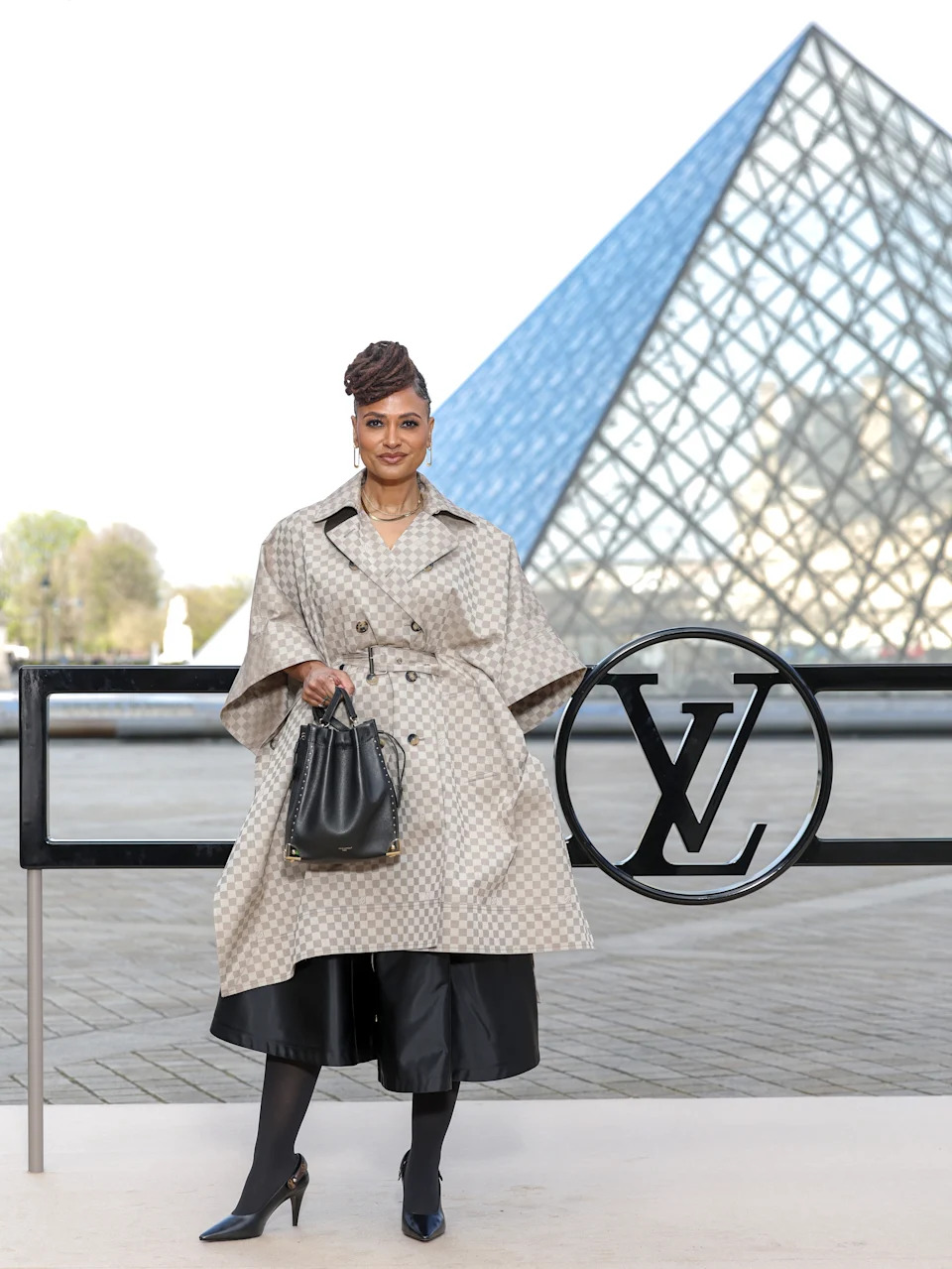 Person in a patterned trench coat and leather pants stands by the Louvre Pyramid, holding a designer bag