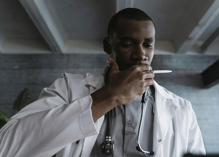 A doctor in a white coat with a stethoscope, thoughtfully holding a pen near his mouth in a dimly lit room.