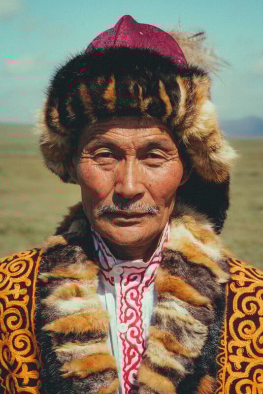 An older man in traditional attire with ornate patterns, wearing a fur hat and coat, stands outdoors in a grassy plain under a blue sky.