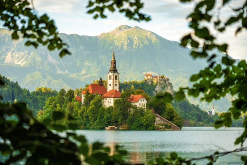 A church with a tall steeple stands on a small island in a lake, surrounded by trees and mountains in the background, viewed through leafy branches.