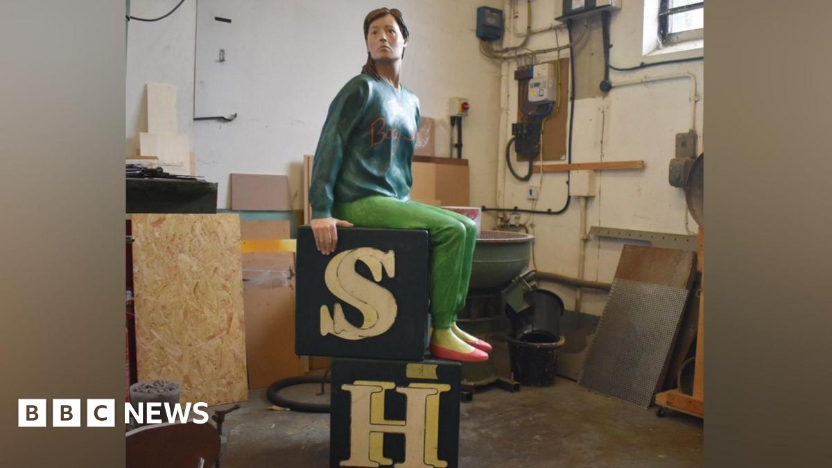 A restored sculpture shows a seated human figure positioned on two stacked black blocks with large cream‑coloured letters “S” and “H.” The sculpture is placed inside a workshop with tools, cables, wooden panels and machinery visible in the background. The figure wears a long‑sleeved top, trousers and shoes.