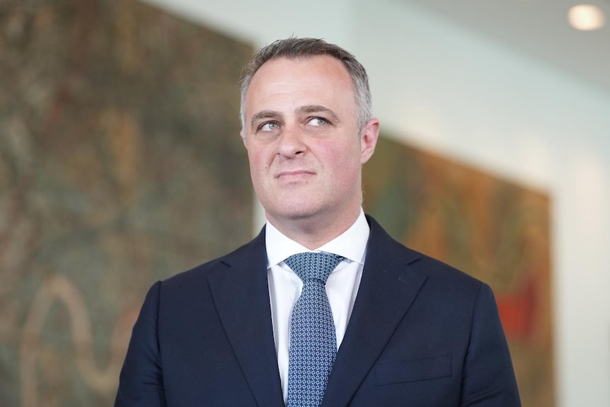 A man with short, greying hair wears a dark suit as he stands in a room with large artworks on the wall.