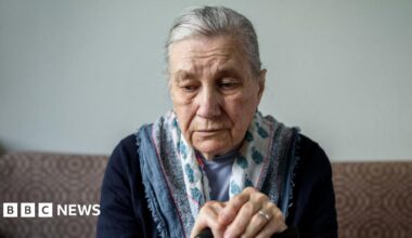 A woman with dark hair and a multiple colour jumper siting on a green sofa inside her living room.