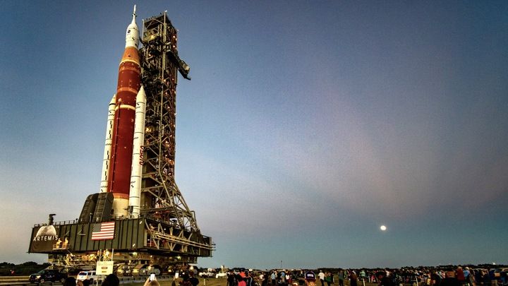 a rocket stands on a launch tower against a fading blue sky and a distant full moon casting rays outward.