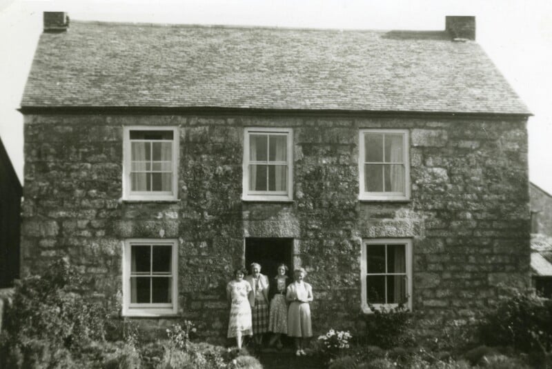 Three people stand in front of an old stone house with four windows and a central door. The house has a pitched roof, and there are plants growing in the garden in front of it. The photo appears to be vintage.