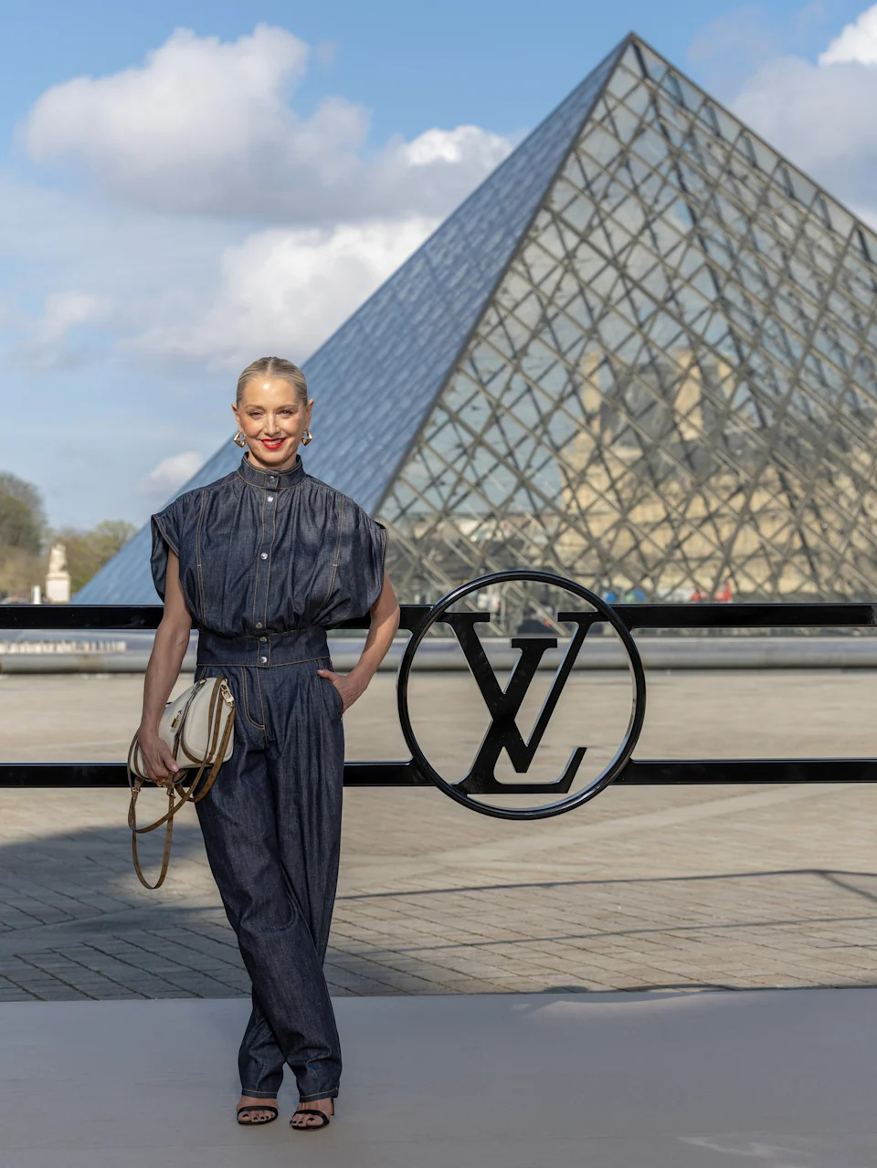 A person in a stylish denim jumpsuit stands in front of the Louvre Pyramid, holding a designer bag