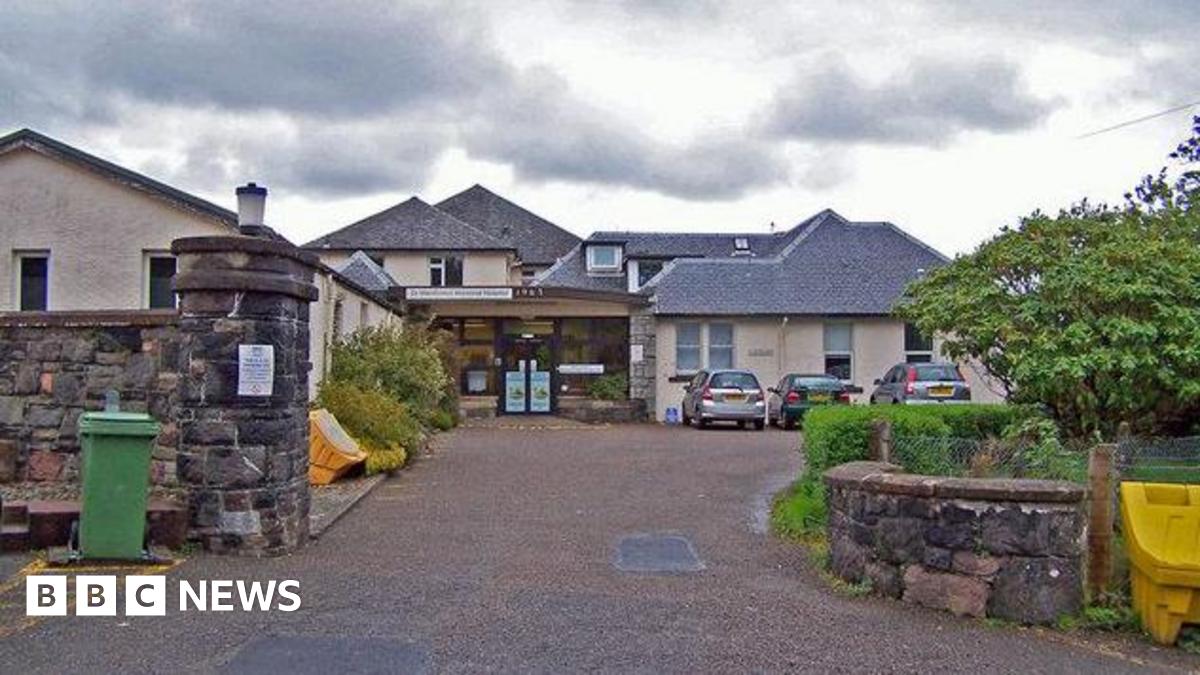 The hospital is made up of a number of low buildings with peaked, slate roofs. Three cars are parked near the glass entrance.