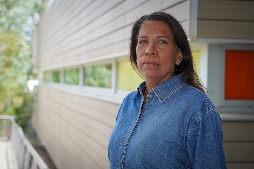A woman with long dark hair wearing a denim shirt stands in front of a light coloured building