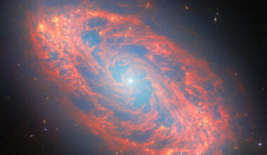 A spiral galaxy, seen tilted diagonally. It has a blue-white, glowing spot at its core. Its oval-shaped disc glows faintly blue throughout with light from its many stars. The disc is filled with waves and strands of bright red dust that swirl around the core. At places there are holes torn in the dust, while elsewhere it forms dense clumps that glow orange. Several tiny, distant galaxies appear across the background.