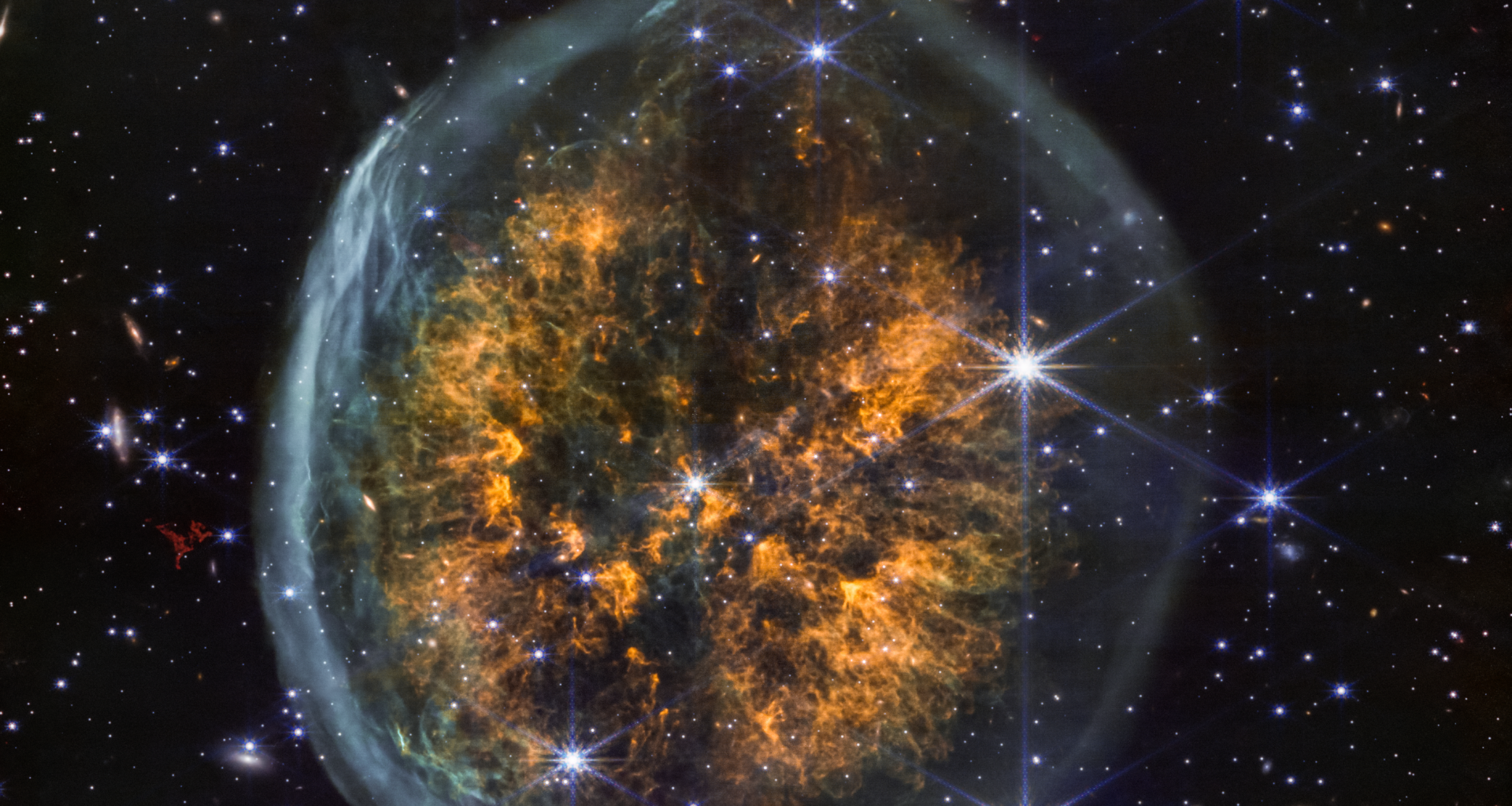 A shell of ghostly gas encapsulates a cloud of amber-colored gases that blow out in both directions from a central point. This makes the nebula look like a top-down view of two brain hemispheres inside a transparent skull. The scene is decorated with multicolor dots of light, representing distant galaxies and stars. The stars shown here have six points, characteristic of Webb images.