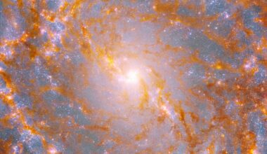A close-up view shows a galaxy’s bright, yellow center, surrounded by patches of glowing orange-brown dust and pink stars.