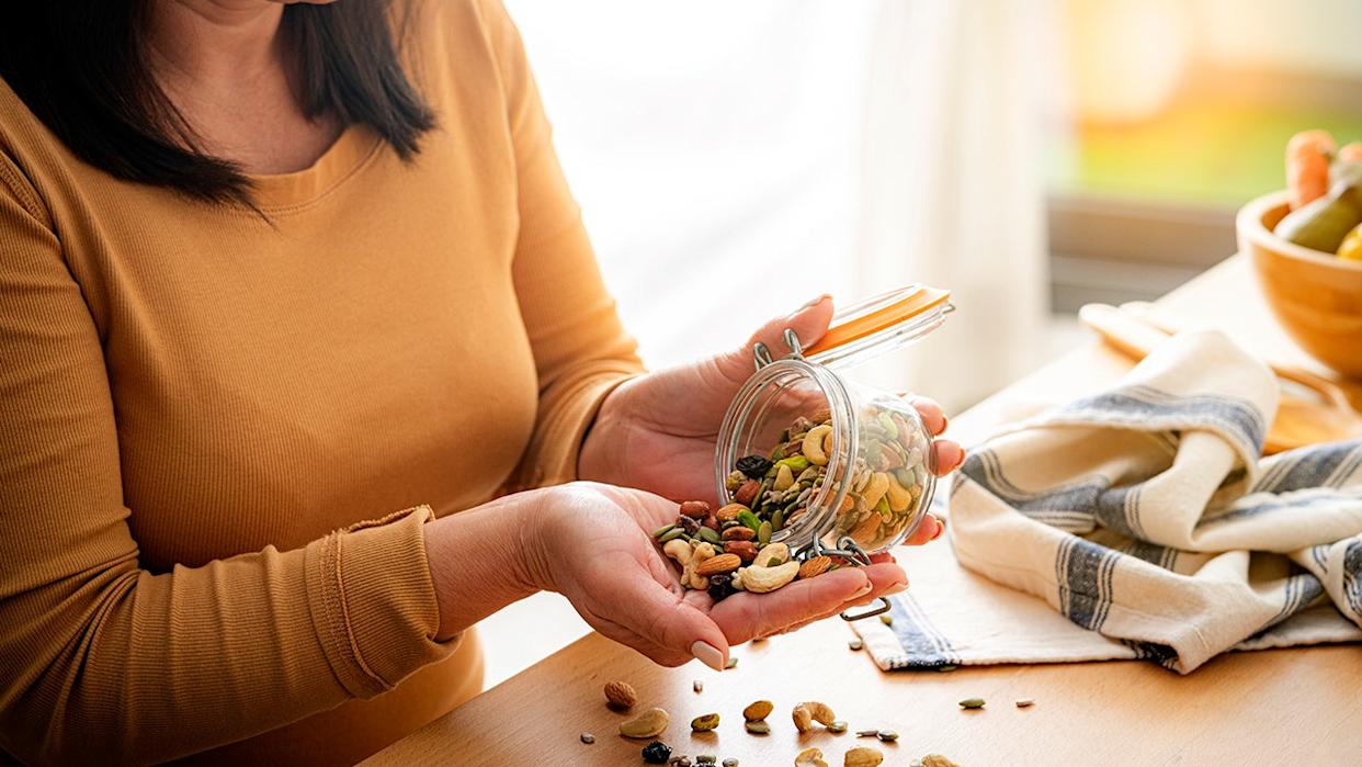 Person pouring mixed nuts and seeds from a glass jar into their hand in a bright kitchen setting.