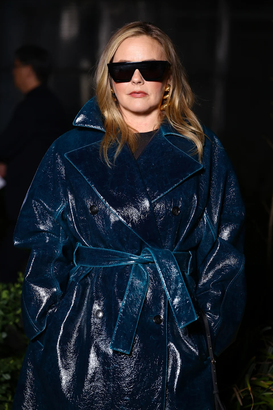 Person in a glossy coat and large sunglasses on a red carpet, exuding a stylish, modern look