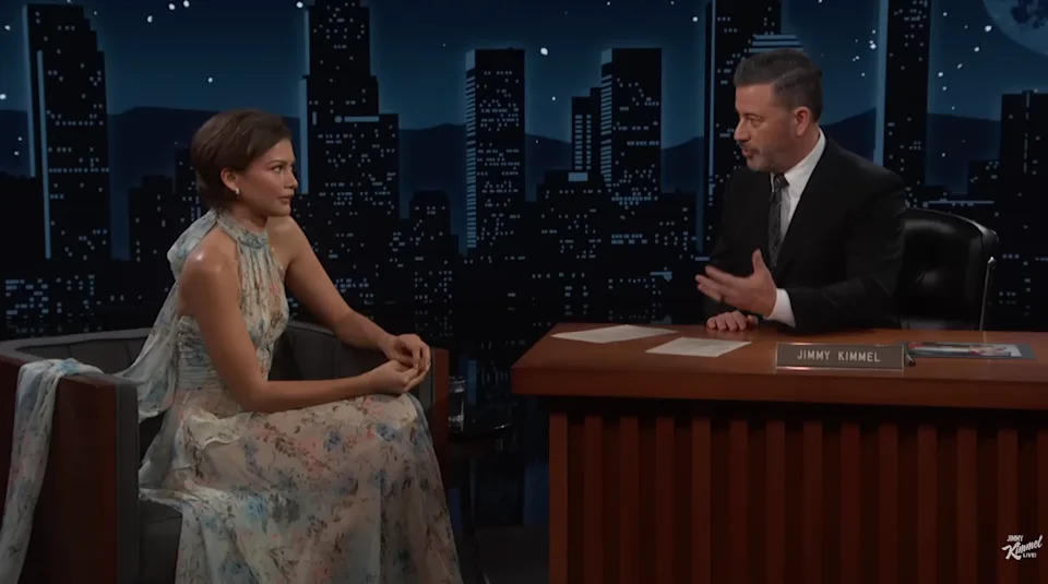 A woman in a flowing, floral gown sits on a talk show set, engaged in conversation with the host who is in a suit