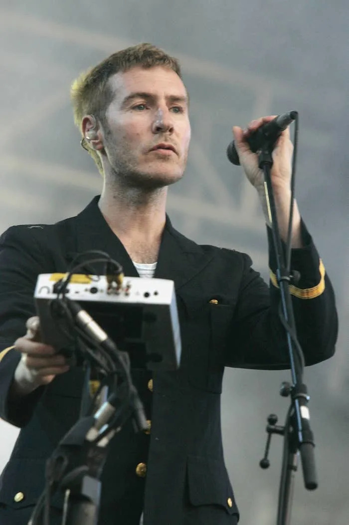 A man in a dark blazer holding a microphone stands in front of audio equipment at an outdoor event