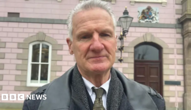 Tom Binet. He has short grey hair and is wearing a jacket, a tweed jacket underneath and a white shirt and dark tie. He is standing in front of a government building.