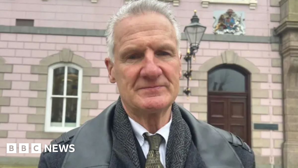 Tom Binet. He has short grey hair and is wearing a jacket, a tweed jacket underneath and a white shirt and dark tie. He is standing in front of a government building.