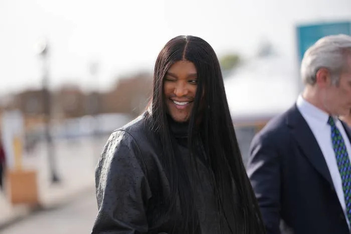 Law Roach smiling, wearing a long coat and having long, straight hair