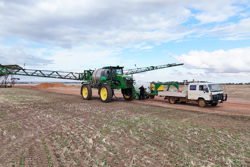 A green sprayer vehicle with yellow tyre rims, parked on a dirt track next to a white ute.